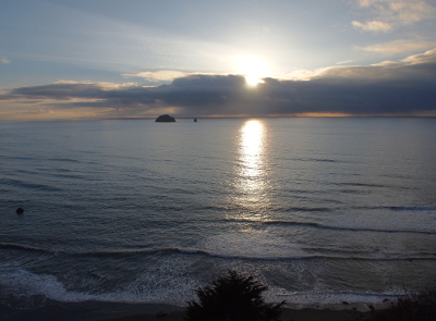 [The setting sun is a glowing orb just above the clouds handing over the ocean. It lights up a portion of the water leading to the foreground as well as the entire blue sky behind and above the clouds.]